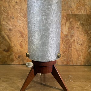 May include: A table lamp featuring a cylindrical, textured, translucent white glass shade secured by two small brass-toned metal knobs. The shade rests on a dark brown wooden base supported by three angled, tapered wooden legs.