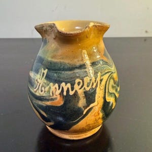 May include: A small ceramic pitcher with a rounded body and a flared rim. The surface features a marbled glaze in shades of mustard yellow, deep blue, and dark green. The word Annecy is embossed in raised, light-colored script across the front.