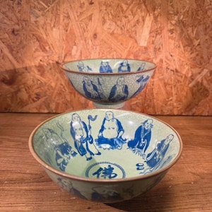 Peut inclure: Deux bols en céramique avec une finition émaillée craquelée vert clair et des personnages peints à la main en bleu représentant des individus assis. L'intérieur du bol avant présente un emblème circulaire central avec le caractère chinois bleu pour Bouddha.