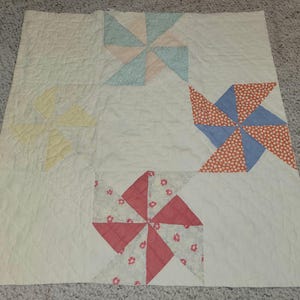 May include: A handmade quilt with a pinwheel design. The quilt features four pinwheel blocks in pastel colors: yellow, blue, pink, and red. The background is a cream color with a quilted pattern.