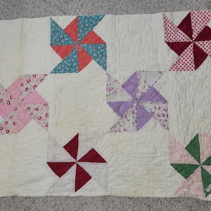 May include: A handmade quilt with a cream-colored background and pinwheel designs. The pinwheels feature various colors and patterns, including red, pink, teal, and purple. The quilt is likely made of cotton and has a quilted texture.