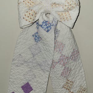 May include: A large decorative bow constructed from quilted fabric featuring a white base with various patches in floral patterns of blue, pink, and yellow, along with solid purple square accents. The quilted texture covers the entire surface.