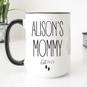 May include: White ceramic mug with a black handle and black rim. The mug has black text that reads "Alison's Mommy Est. 2023" with two small black footprints below the text.