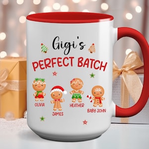 May include: White ceramic mug with a red rim and handle. The mug features a gingerbread person design with the text "Gigi's Perfect Batch" and the names "Olivia", "James", "Heather", and "Baby John".