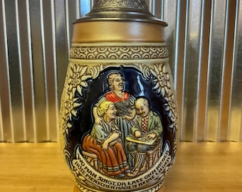 May include: A ceramic beer stein with a detailed relief scene of people singing, framed by floral and geometric patterns. The stein has a metal lid and a handle. The base and lid are brown, with a tan body and blue accents.