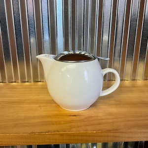 May include: A white ceramic teapot with a rounded body, a curved handle, and a spout. It has a brown lid with a silver rim and a small infuser. The teapot is sitting on a wooden surface.