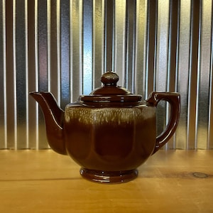 May include: A brown ceramic teapot with a white glaze. The teapot has a rounded body and a spout on the side. The teapot is sitting on a wooden surface.