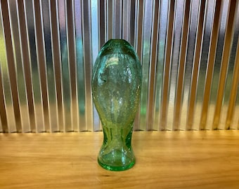 May include: A tall, green glass vase with a crackled texture. The vase has a flared base and a narrow neck, standing on a wooden surface against a corrugated metal background. The vase is empty.