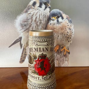 May include: A vintage ceramic beer stein with a beige rim and base, featuring a red crest with a black lion and the text "Bohemian Beer Detroit, Mich. U.S." The stein is decorated with ornate patterns and is set against a backdrop of two birds.
