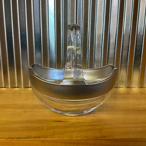 May include: A clear glass and silver-toned serving dish with a handle. The dish has a rounded shape with a silver band around the middle. The handle is clear and rectangular. The dish is sitting on a wooden surface.