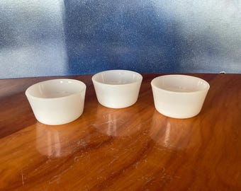 May include: Three small, white, ceramic bowls with a slightly flared shape. The bowls are arranged on a wooden surface, reflecting the warm tones of the wood. The background is a gradient of blue and white.