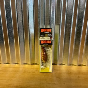 May include: A Rapala Fat Rap fishing lure in its original packaging. The lure is brown and yellow with a reflective finish and two treble hooks. The packaging has the Rapala logo and product information.