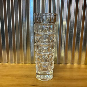 May include: A clear glass vase with a geometric design. The vase is cylindrical with a series of stacked, diamond-shaped patterns. The vase is sitting on a wooden surface, with a corrugated metal background.