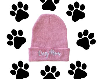 May include: Pink knit beanie with a white embroidered "Dog Mom" on the cuff. The beanie is surrounded by black paw prints.