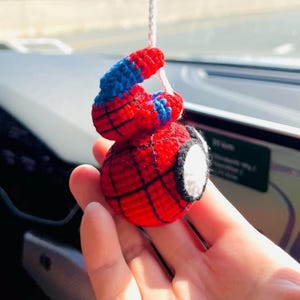 May include: A handmade crochet Spider-Man car charm. The charm is red with black web lines, a blue and red head, and a white eye. It hangs from a white string. The charm is held in a hand.