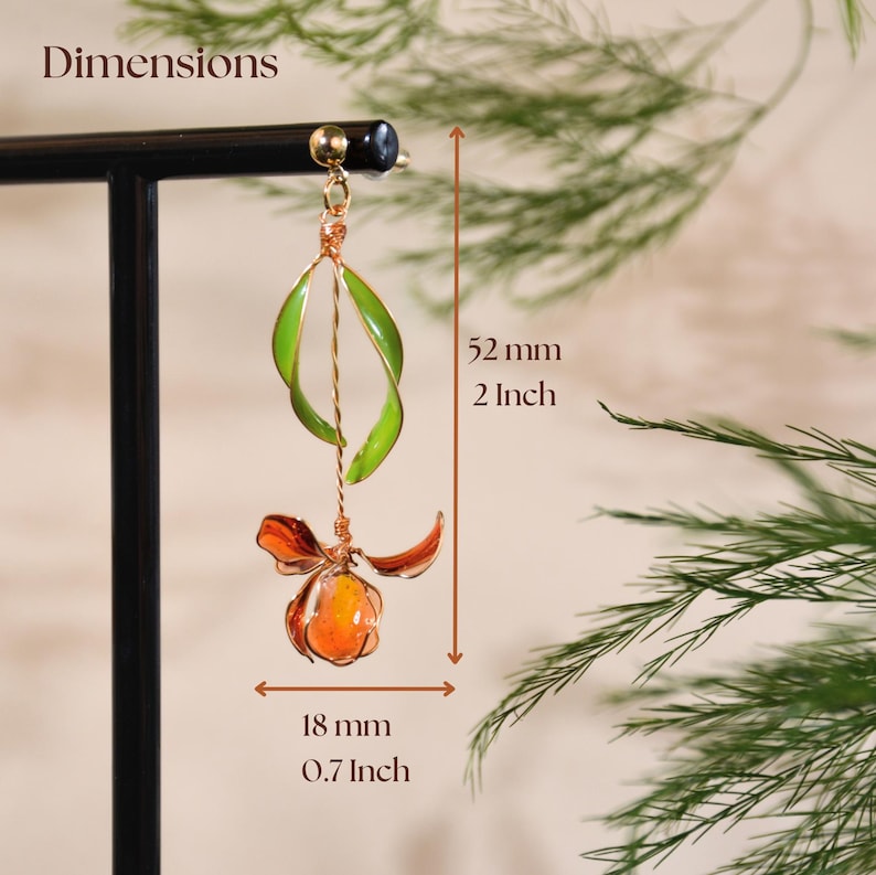 May include: A dangling earring featuring a green leaf-shaped element and an orange flower-shaped element connected by a thin wire. The earring measures 52 mm or 2 inches in height and 18 mm or 0.7 inches in width, as indicated by the text Dimensions.
