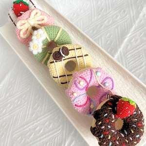 Op de afbeelding: Vijf gehaakte donuts op een wit bord. De donuts zijn versierd met verschillende toppings, waaronder chocolade, sprinkles en aardbeien.