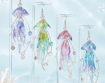 Puede incluir: Cuatro pendientes colgantes con forma de medusa, cada uno con pétalos de resina translúcida multicolor y ganchos plateados. Los pendientes muestran degradados en morado, verde, rosa y azul, acentuados con pequeñas cuentas iridiscentes.