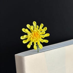 May include: A yellow crocheted flower bookmark with a green stem. The bookmark is placed in the corner of a white book.