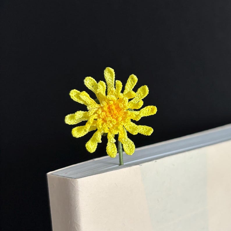 May include: A yellow crocheted flower bookmark with a green stem. The bookmark is placed in the corner of a white book.