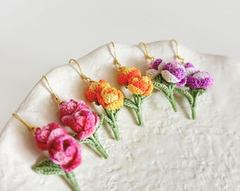 Puede incluir: Una colección de pendientes de flores de ganchillo hechos a mano en varios colores, incluyendo rosa, naranja, amarillo y morado. Cada pendiente presenta un diseño floral detallado con tallos y hojas verdes, y ganchos dorados. Los pendientes se exhiben sobre una superficie blanca texturizada.