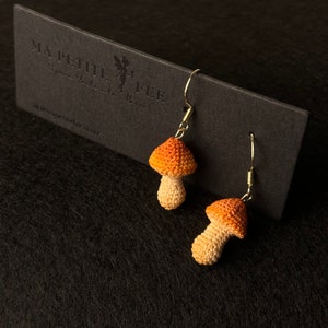 May include: A pair of crocheted mushroom earrings with orange and beige accents. The earrings are hanging from gold hooks.