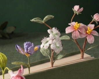 Puede incluir: Un conjunto de cuatro marcapáginas de ganchillo con forma de flor insertados en un libro verde. Las flores incluyen una fucsia rosa, un iris morado, un racimo de flores blancas con hojas verdes y una flor rosa con estambres amarillos.