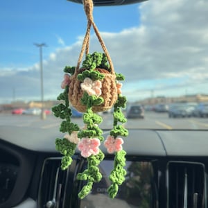 May include: A crocheted hanging plant with green leaves and pink flowers. The plant is in a brown pot and is hanging from a rearview mirror in a car.