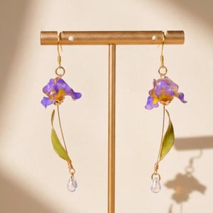 Könnte beinhalten: Ein Paar goldfarbene Ohrhänger mit violetten und gelben Irisblüten-Anhängern sowie grünen Stielen und Blättern. Jeder Ohrring ist mit einer kleinen, irisierenden tropfenförmigen Perle versehen, die am unteren Ende des Stiels hängt.
