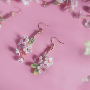 May include: Delicate floral earrings featuring clusters of pink and white blossoms with green leaves. The earrings have gold-colored hooks and are set against a soft pink background. These earrings are a charming accessory for spring.