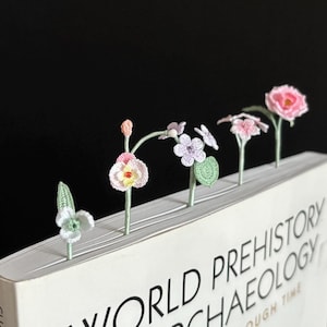 May include: Five crocheted flower bookmarks in pink, white, and yellow are placed in the pages of a book titled "World Prehistory Archaeology".