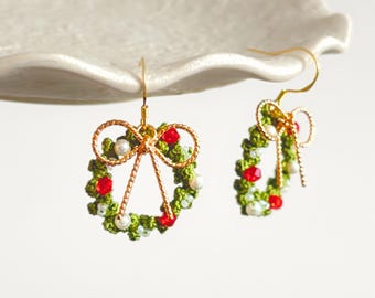 May include: A pair of Christmas wreath earrings. Each earring features a green crocheted wreath adorned with red beads and white pearls. A gold-colored bow and hook complete the design, creating a festive and delicate accessory.