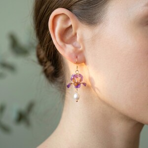 May include: A gold-toned earring featuring a purple and yellow flower-shaped charm suspended from a thin wire hook. Below the flower, a single white spherical pearl hangs from a small gold-toned link, creating a delicate drop design.