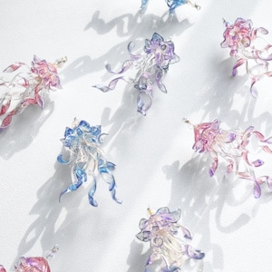 Pode incluir: Uma coleção de ornamentos de água-viva de vidro iridescente em tons de rosa, azul e roxo. As águas-vivas são suspensas por um fio e têm tentáculos delicados e fluidos.