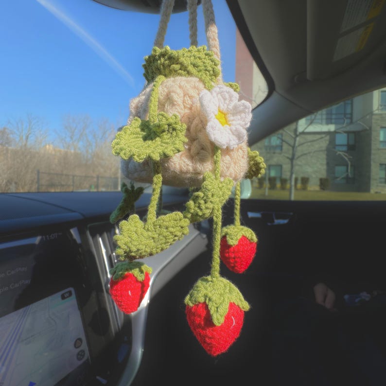 May include: A handmade crochet car charm featuring a beige pouch with green leaves and a white flower. Three red crochet strawberries hang below. The charm is suspended from the car's rearview mirror.