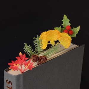 Può includere: Un segnalibro all'uncinetto con una composizione botanica di fiori rossi, fronde di felce verdi, pigne marroni, foglie di ginkgo gialle e foglie di agrifoglio verdi con bacche rosse, inserito nella parte superiore di un libro grigio con testo.
