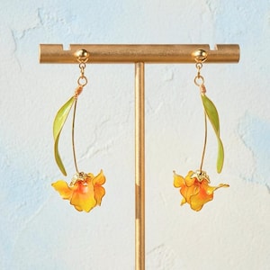 May include: A pair of drop earrings featuring orange and yellow flower blossoms with green leaves attached to thin gold-toned stems. The earrings are suspended from gold-toned ball-post studs, showcasing a delicate floral design with vibrant petals.