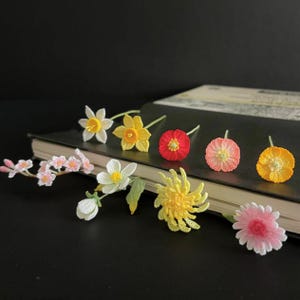 May include: A collection of handmade crocheted flowers in various colors, including yellow, pink, red, and white. The flowers are arranged on a dark surface and appear to be delicate and intricate.
