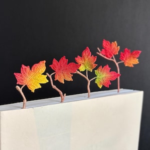 May include: Five crocheted maple leaves in shades of red, orange, and yellow are attached to a white book. The leaves are arranged in a row, with the stems pointing towards the top of the book.