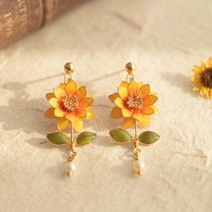 May include: A pair of gold-tone stud earrings featuring a yellow and orange sunflower design with green leaves on a thin stem and a small white pearl dangling at the bottom. The petals have a gradient color effect and the center is textured.