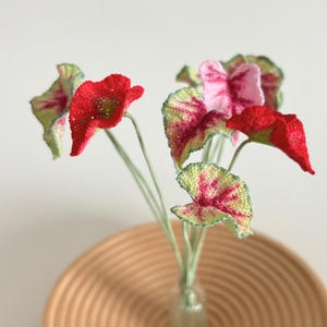 May include: A bouquet of handmade fabric flowers in a clear glass vase. The flowers feature red, pink, and green leaves with intricate details. The arrangement sits atop a woven, light brown basket.