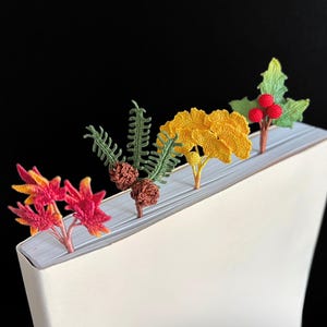 Puede incluir: Cinco marcapáginas de ganchillo con forma de diferentes plantas. Los marcapáginas son rojos, amarillos, verdes y marrones. Están colocados en el borde de un libro blanco.