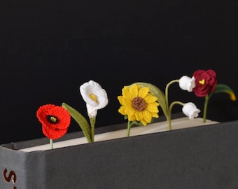 Puede incluir: Un conjunto de cinco marcapáginas de flores de ganchillo insertados en un libro gris. Las flores incluyen una amapola roja, un lirio de agua blanco, un girasol amarillo, lirios de los valles blancos y una rosa roja oscura, con tallos verdes.
