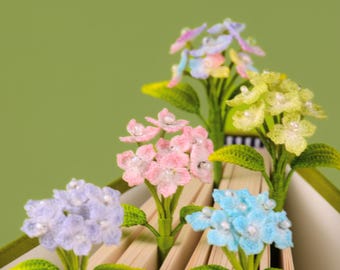 Puede incluir: Cinco marcapáginas de flores de ganchillo con racimos similares a hortensias en tonos pastel de lavanda, rosa, amarillo y azul. Cada flor está acentuada con pequeñas cuentas blancas en el centro y unida a un tallo verde, insertada en las páginas.