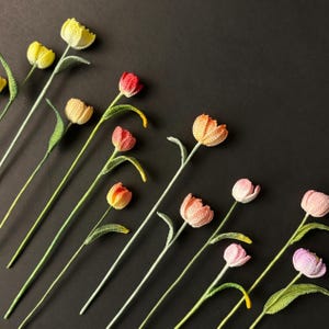 Puede incluir: Flores de tulipán tejidas a ganchillo en varios colores, incluyendo amarillo, naranja, rojo y rosa, dispuestas diagonalmente sobre un fondo negro. Cada flor tiene un tallo largo y hojas verdes, mostrando el detalle artesanal.