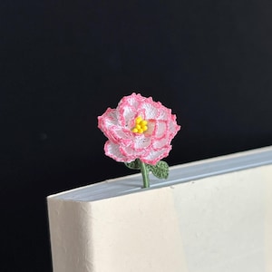 May include: A pink and white crocheted flower bookmark with a yellow center. The bookmark is placed in the corner of a white book.