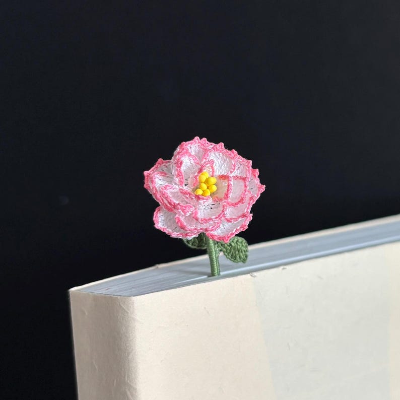 May include: A pink and white crocheted flower bookmark with a yellow center. The bookmark is placed in the corner of a white book.