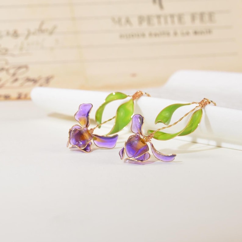 May include: A pair of earrings featuring translucent purple and orange flower petals with bright green leaves, crafted with thin gold-toned wire stems. The floral design displays delicate, curved shapes and vibrant colors, capturing the essence of a blooming plant.