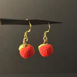 May include: A pair of handmade crochet apple earrings. Each earring features a small, red apple with a green and yellow leaf and a gold-colored hook. The earrings are suspended against a dark background.