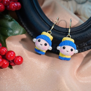 May include: A pair of crocheted earrings featuring a smiling face with blue and yellow accents. The earrings are attached to gold hooks.